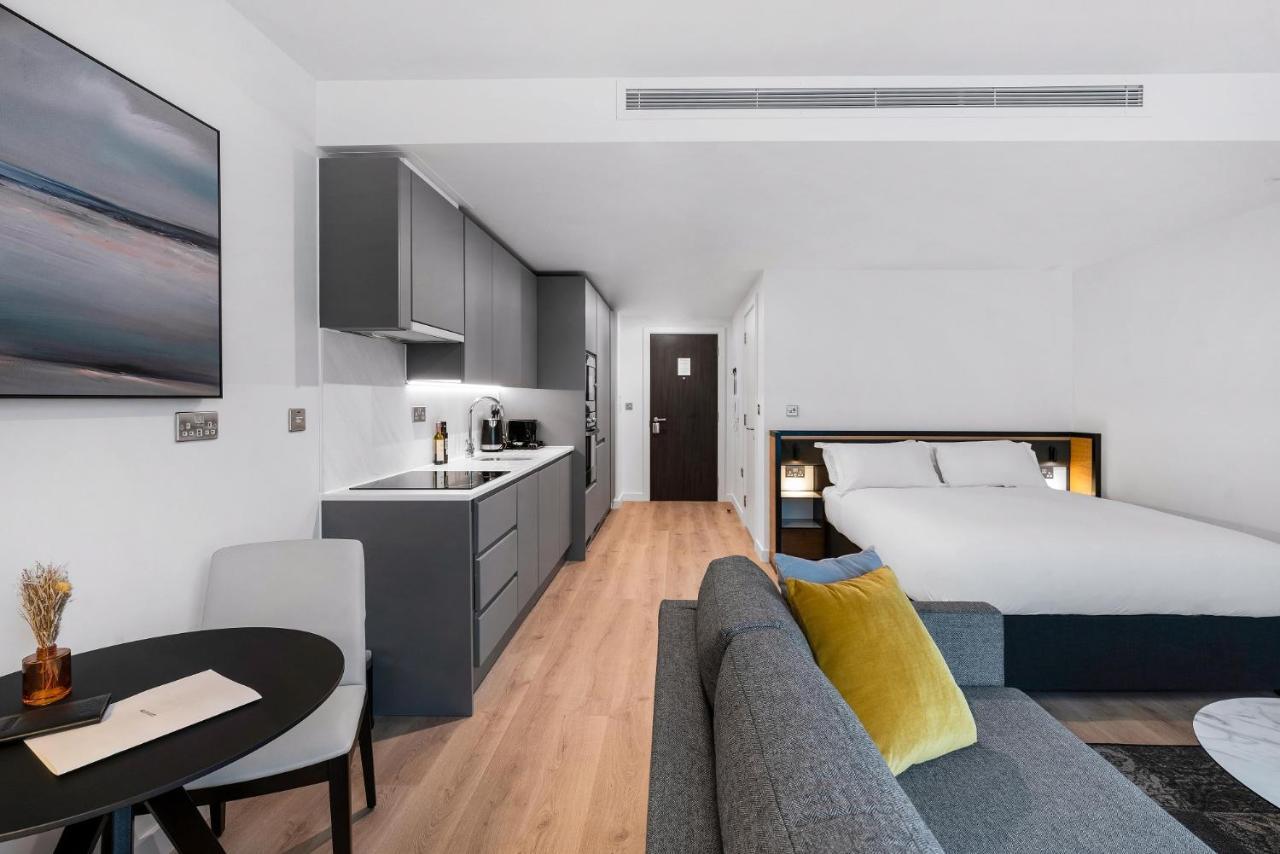 Citysuites 2 Aparthotel Σάλφορντ Εξωτερικό φωτογραφία The photo shows a modern and stylish studio apartment. It features a kitchenette with dark cabinetry and a small dining table with two chairs. There's a cozy sofa with colorful cushions, positioned next to the table. In the background, a large bed wi