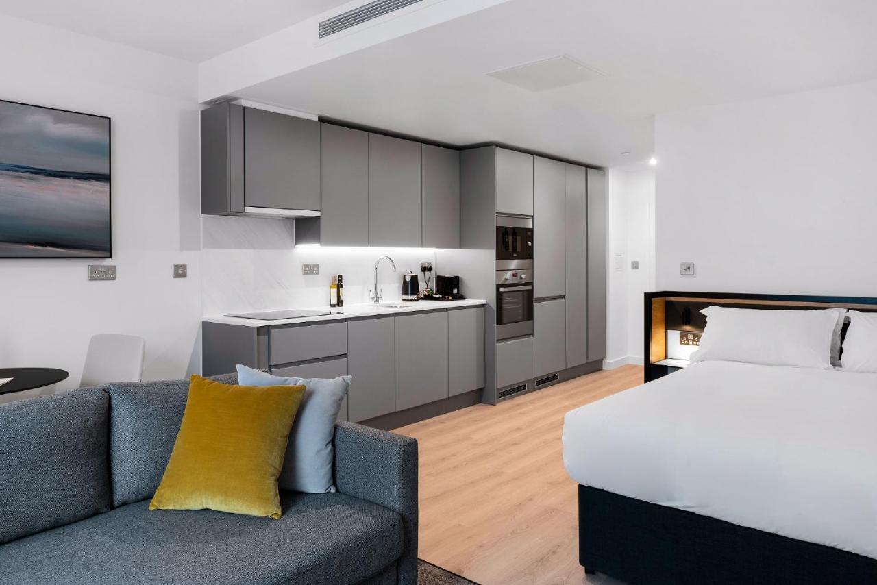 Citysuites 2 Aparthotel Μάντσεστερ Εξωτερικό φωτογραφία The photo shows a modern studio apartment featuring an open-plan layout. On one side, there is a kitchenette with sleek, gray cabinets and appliances, including a coffee maker and oven. Adjacent to the kitchenette is a small dining area with a table 