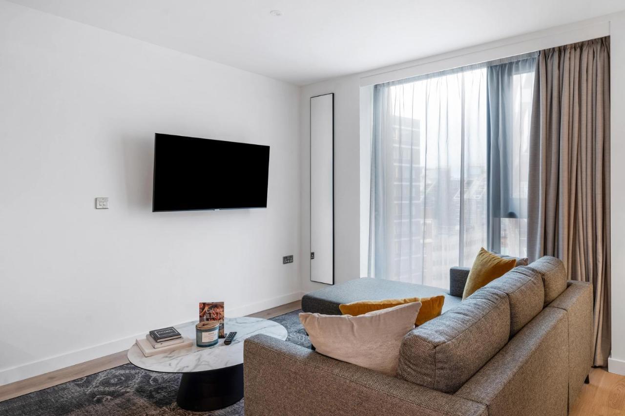 Citysuites 2 Aparthotel Μάντσεστερ Εξωτερικό φωτογραφία The image shows a modern living room with a minimalist design. There is a large flat-screen TV mounted on the white wall. A comfortable gray sofa with textured cushions occupies the center of the room, complemented by a round coffee table in front of