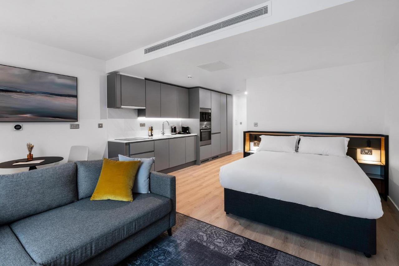 Citysuites 2 Aparthotel Σάλφορντ Εξωτερικό φωτογραφία The photo shows a modern studio apartment. On one side, there is a comfortable gray sofa adorned with a yellow cushion. Nearby, a round table is placed, likely intended for dining or casual sitting. 

The kitchen area features sleek gray cabinetry wi