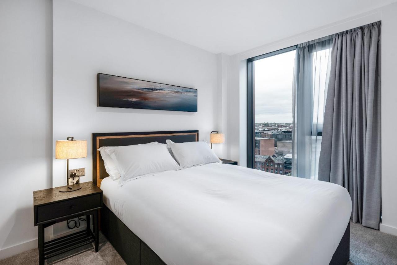Citysuites 2 Aparthotel Μάντσεστερ Εξωτερικό φωτογραφία The image shows a modern bedroom featuring a large bed with a white duvet and two pillows. On either side of the bed are small nightstands with lamps. A large window occupies one wall, draped with light gray curtains, allowing natural light to enter 