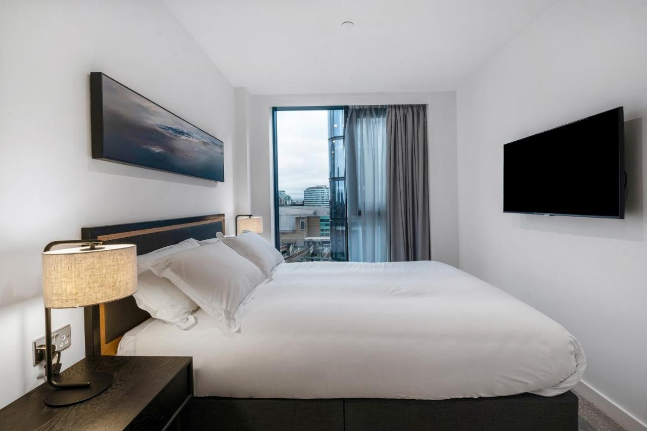 Citysuites 2 Aparthotel Σάλφορντ Εξωτερικό φωτογραφία The photo shows a modern bedroom with a minimalist aesthetic. It features a large bed with white bedding and two pillows. On either side of the bed, there are bedside tables with lamps. A large window lets in natural light, covered by sheer curtains.