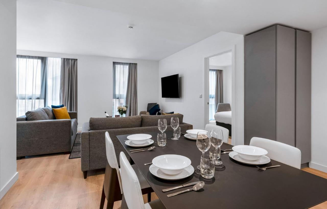 Citysuites 2 Aparthotel Σάλφορντ Εξωτερικό φωτογραφία The photo shows a modern living space with an open layout. In the foreground, there is a dining table set for a meal, featuring multiple plates, glasses, and cutlery arranged neatly. In the background, there is a comfortable-looking gray sofa with a 