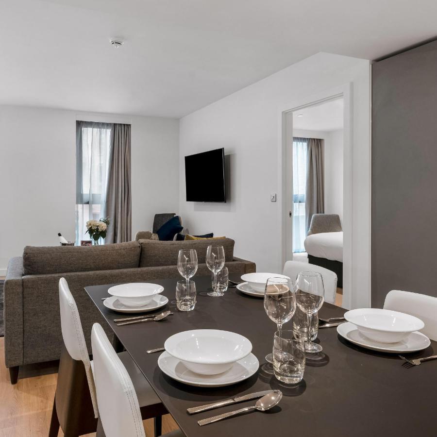 Citysuites 2 Aparthotel Σάλφορντ Εξωτερικό φωτογραφία The photo shows a modern, well-lit living space that features a dining area and a sitting area. In the foreground, there is a dining table set for six, with white plates and crystal glasses arranged neatly. The table is surrounded by white chairs. 

