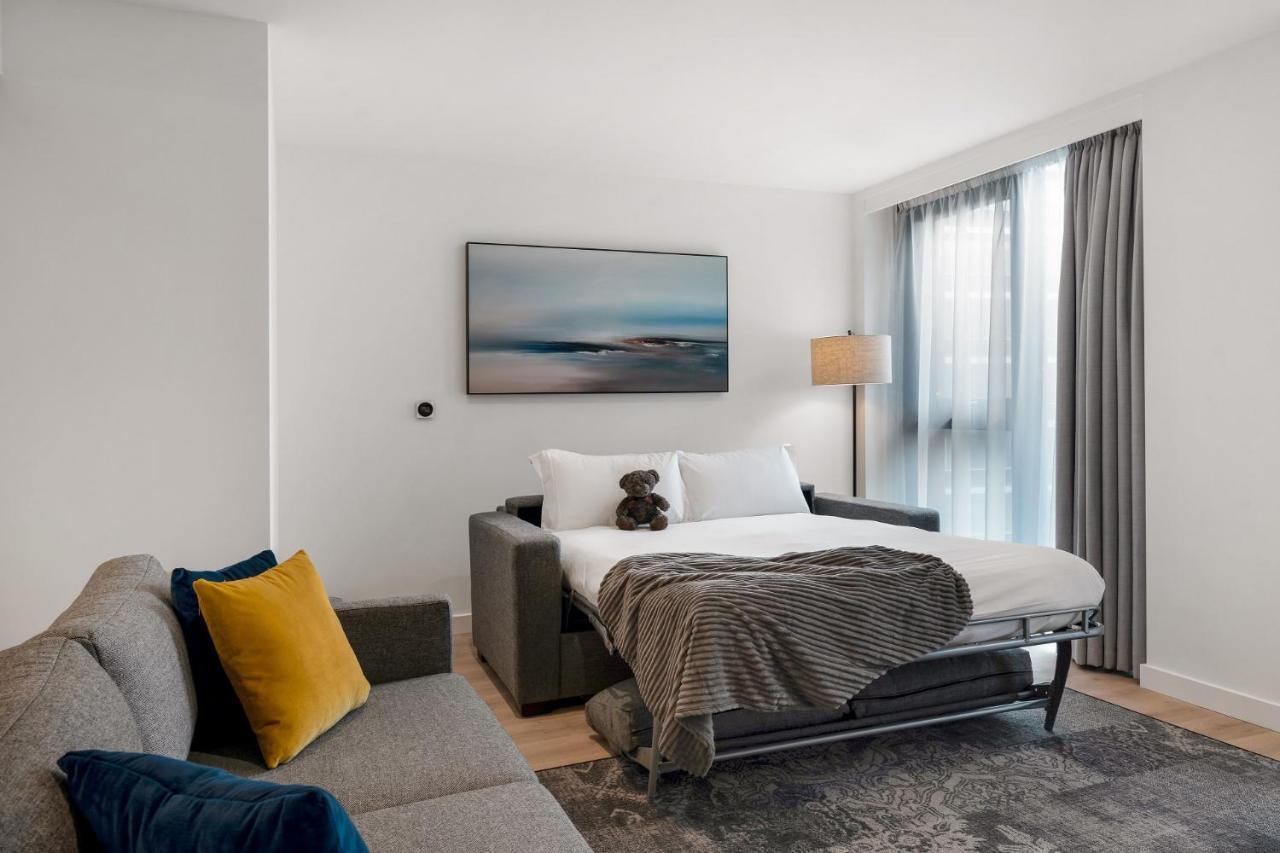 Citysuites 2 Aparthotel Σάλφορντ Εξωτερικό φωτογραφία The photo shows a modern and minimalistic guest room. There is a sofa on the left side, adorned with a colorful cushion. In the middle is a bed that appears to be a pull-out style, featuring white bedding and a gray throw blanket. A stuffed toy is pl