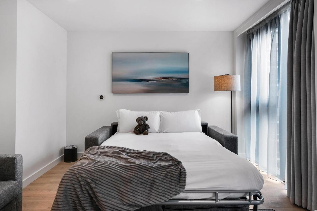 Citysuites 2 Aparthotel Μάντσεστερ Εξωτερικό φωτογραφία The photo shows a modern bedroom featuring a bed with white bedding and a decorative throw blanket. There is a teddy bear placed on the bed. A piece of abstract art is hanging on the wall above the bed. The room has a large window with sheer curtains