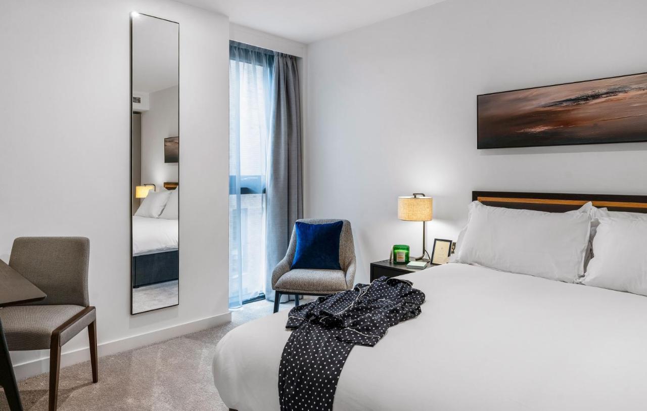 Citysuites 2 Aparthotel Σάλφορντ Εξωτερικό φωτογραφία The photo shows a modern hotel room. It features a neatly made bed with white linens and a dark-colored throw draped over it. There is a small bedside table with a lamp next to the bed. A comfortable chair in a light color is positioned near the wind