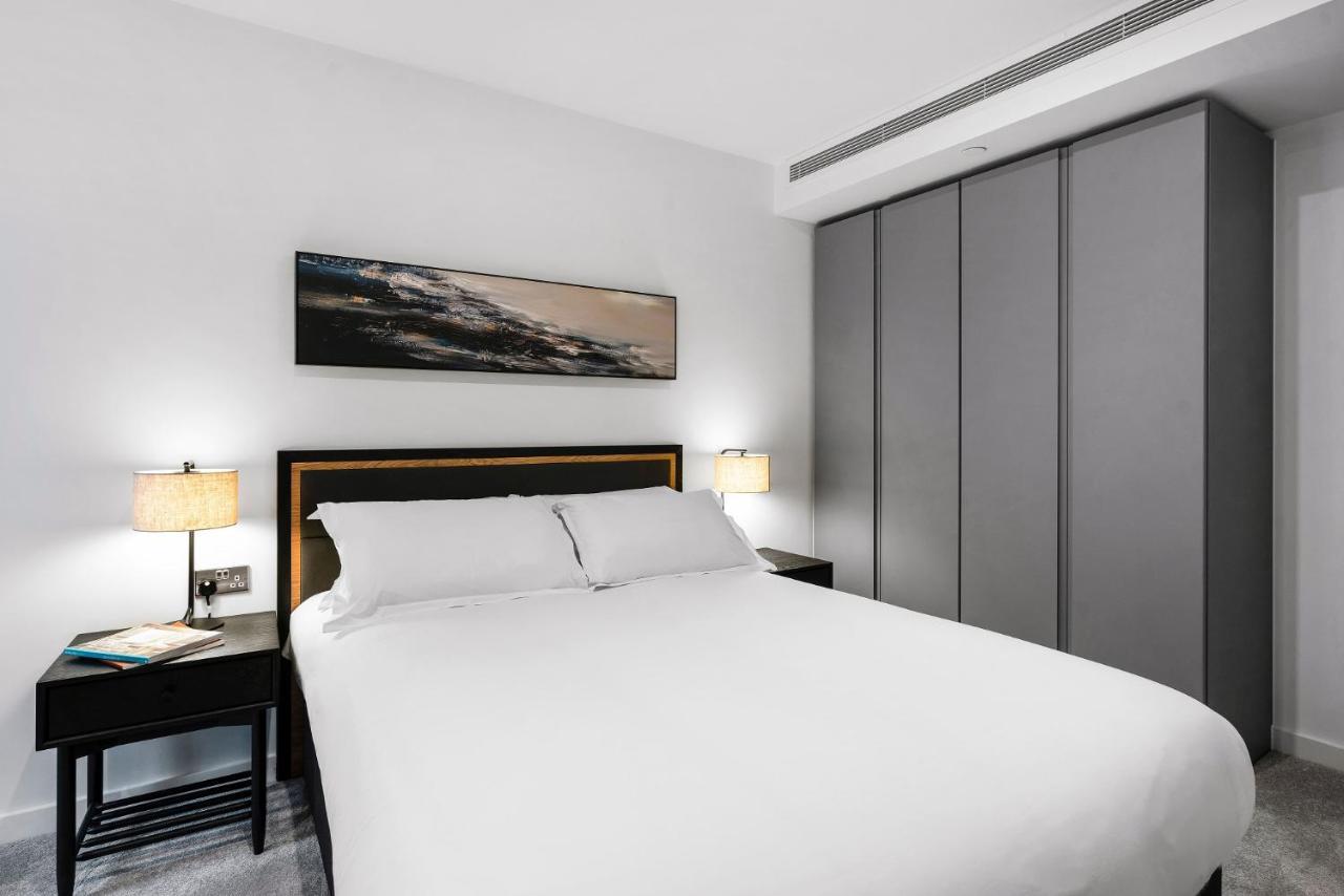 Citysuites 2 Aparthotel Μάντσεστερ Εξωτερικό φωτογραφία The photo shows a modern bedroom designed with a minimalist aesthetic. The focal point is a large bed with white bedding, positioned against a wall. On either side of the bed, there are black nightstands topped with lamps, providing a warm glow. Abov