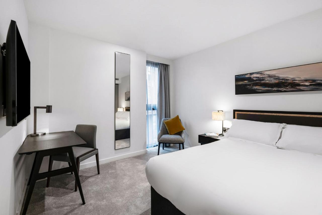 Citysuites 2 Aparthotel Μάντσεστερ Εξωτερικό φωτογραφία The photo shows a modern hotel room with a clean and minimalist design. It features a large bed with white linens, a small desk with a chair, and a mirror mounted on the wall. There is a chair in one corner with a decorative pillow, and the room has 
