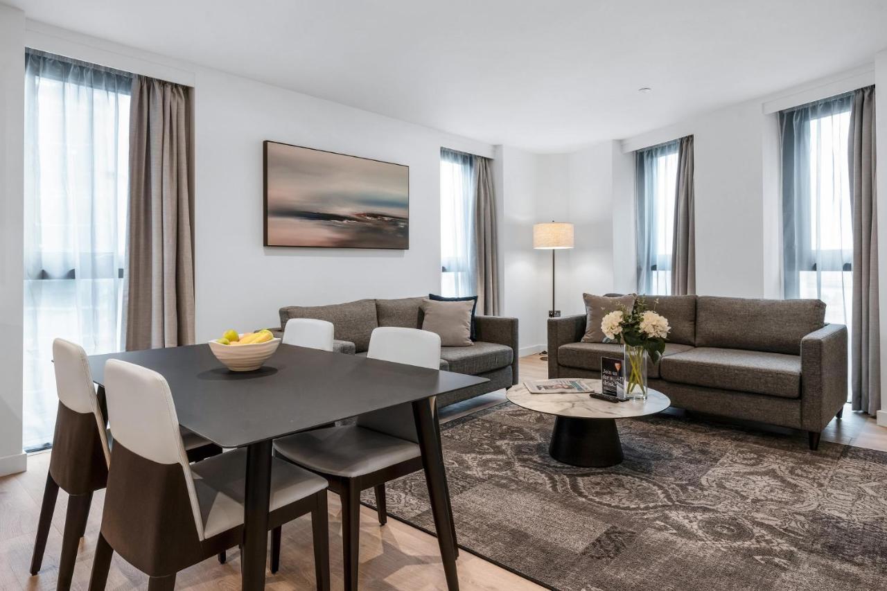 Citysuites 2 Aparthotel Μάντσεστερ Εξωτερικό φωτογραφία The photo depicts a modern and stylish interior of a living space. On the left, there is a dining table with four chairs, featuring a bowl of fruits on the table. In the background, there's a cozy seating area with a gray sofa and a smaller round cof