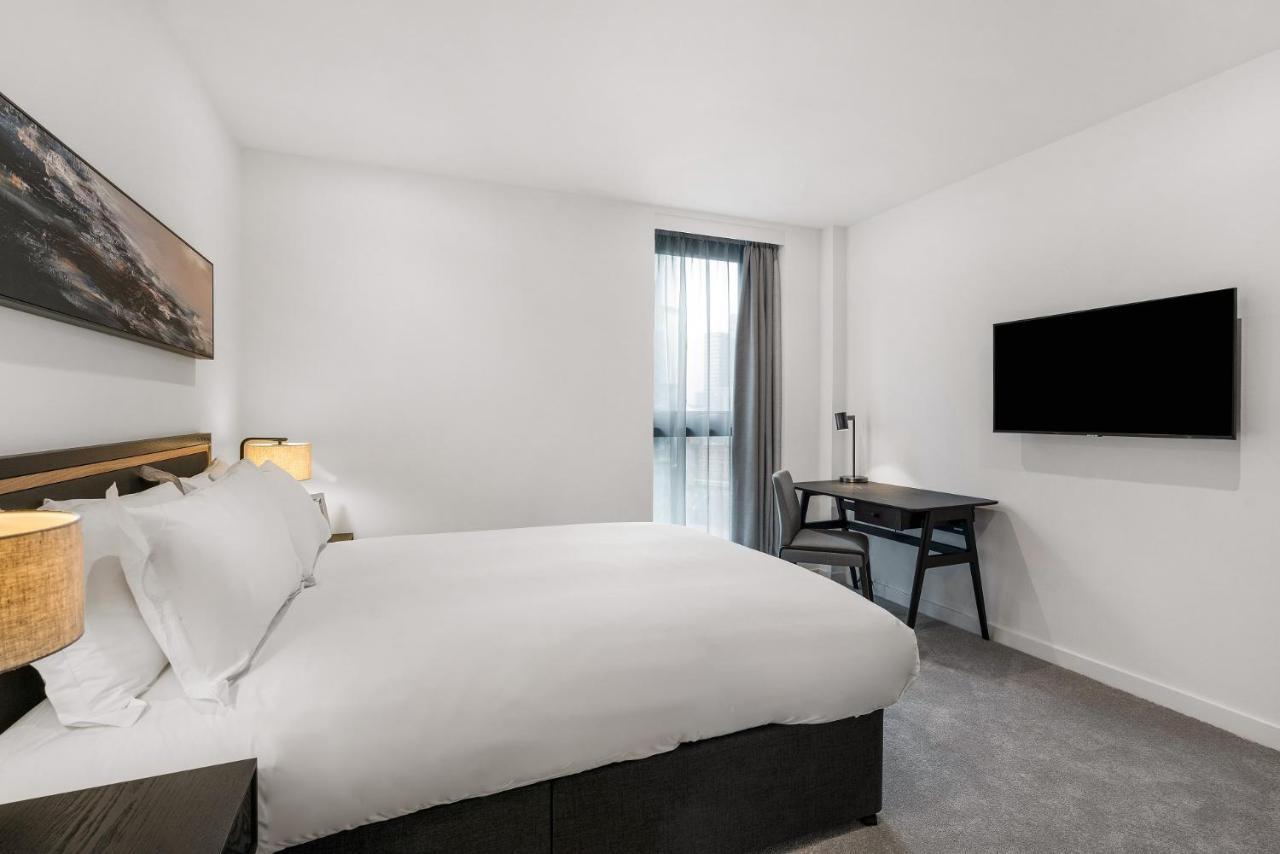 Citysuites 2 Aparthotel Μάντσεστερ Εξωτερικό φωτογραφία The photo shows a modern hotel room featuring a large bed with white linens and a dark headboard. There are two bedside lamps on either side of the bed. A desk with a chair is placed against one wall, and a flat-screen TV is mounted on another wall. 