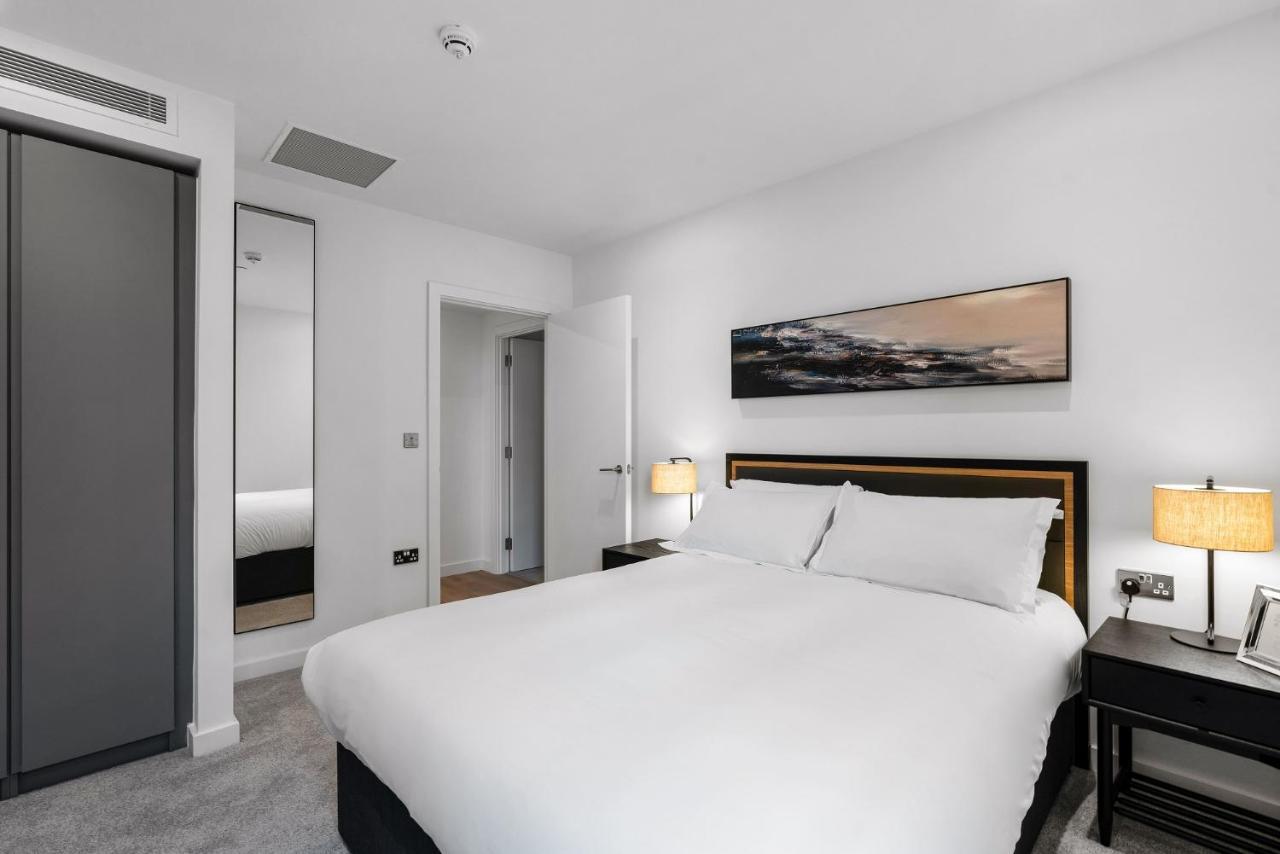 Citysuites 2 Aparthotel Σάλφορντ Εξωτερικό φωτογραφία The photo shows a modern bedroom featuring a large double bed with white bedding. There are two bedside tables with lamps on either side of the bed. The walls are painted white, and there's a piece of artwork above the bed that depicts a landscape. A