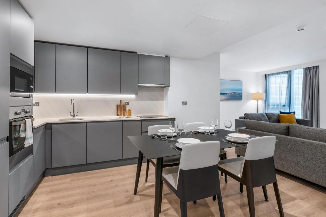 Citysuites 2 Aparthotel Μάντσεστερ Εξωτερικό φωτογραφία The photo shows a modern, open-concept kitchen and living area. The kitchen features sleek, gray cabinetry with a countertop, a sink, and built-in appliances. There are cooking utensils displayed on the countertop. Next to the kitchen, there is a din