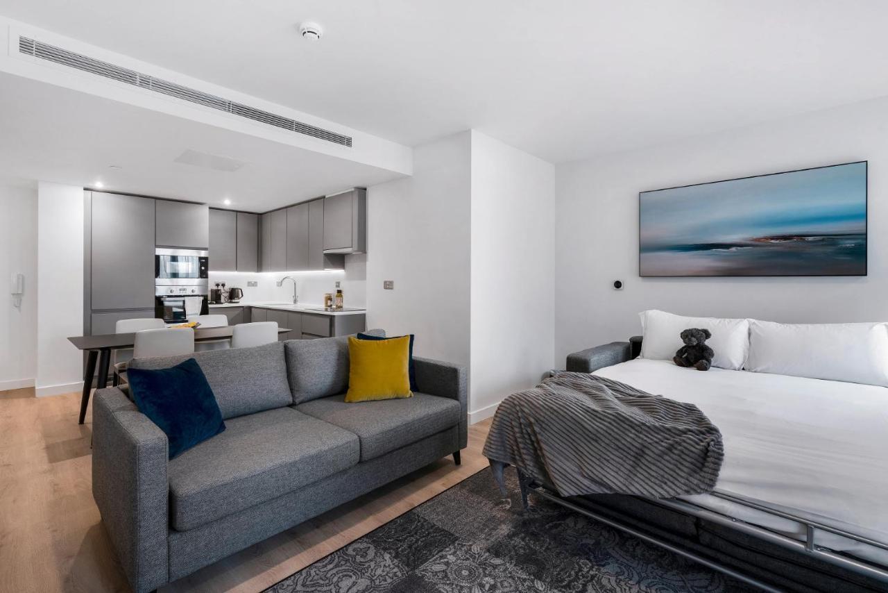 Citysuites 2 Aparthotel Μάντσεστερ Εξωτερικό φωτογραφία The photo shows a modern, open-concept living space that includes a kitchen and a sleeping area. 

- **Living Area**: On the left, there's a gray sofa with a blue and yellow throw pillow. A blanket is draped over one side, and there's a small dark ru