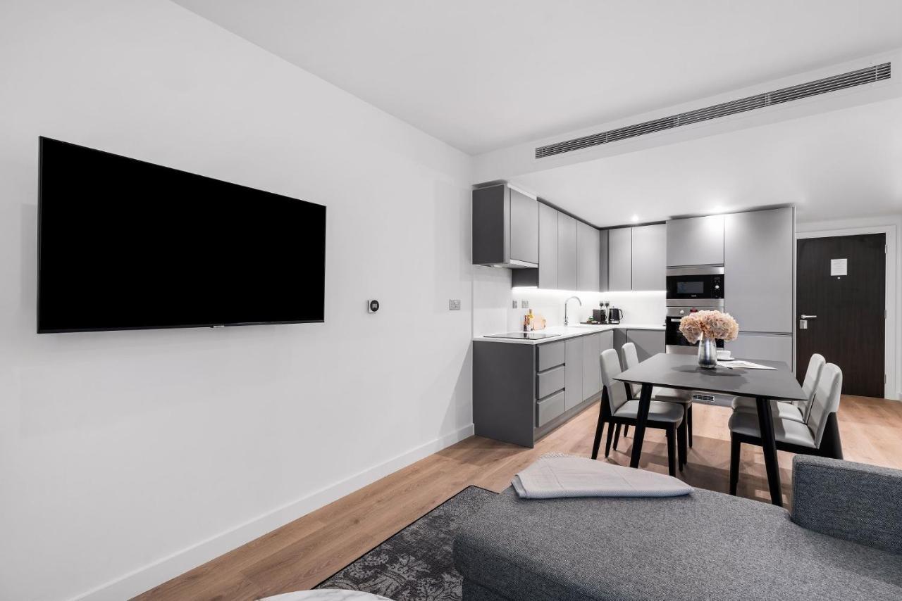 Citysuites 2 Aparthotel Μάντσεστερ Εξωτερικό φωτογραφία The photo shows a modern, minimalist living space. On the left, there is a large flat-screen TV mounted on the wall. To the right, a kitchen area features sleek gray cabinets and appliances, including a built-in microwave. In front of the kitchen, th