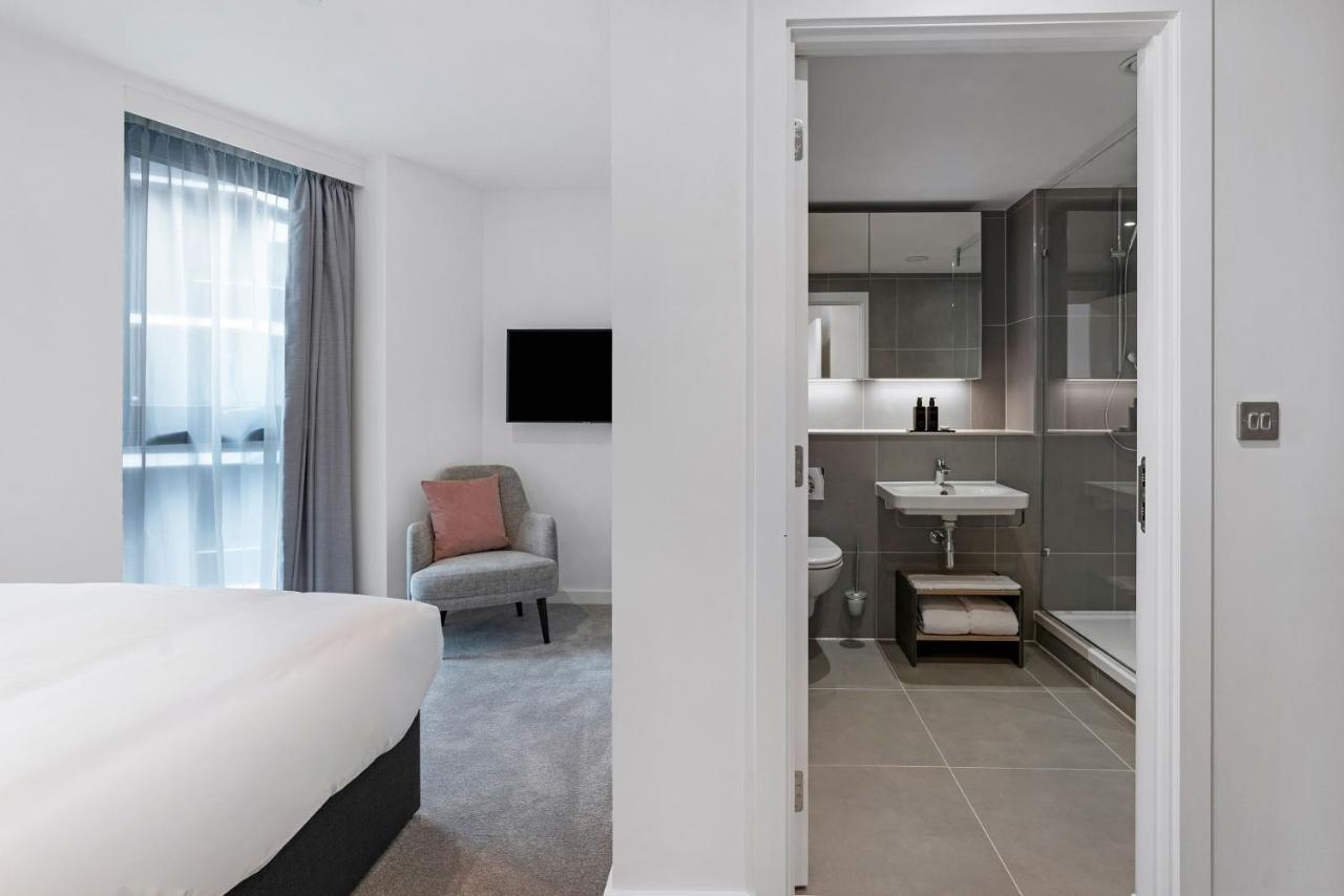 Citysuites 2 Aparthotel Μάντσεστερ Εξωτερικό φωτογραφία The photo shows a modern hotel room and bathroom setup. On the left, there is a bed with white bedding and a small sitting area that includes a gray chair and a pillow in a subtle pink shade. A flat-screen TV is mounted on the wall, providing enterta