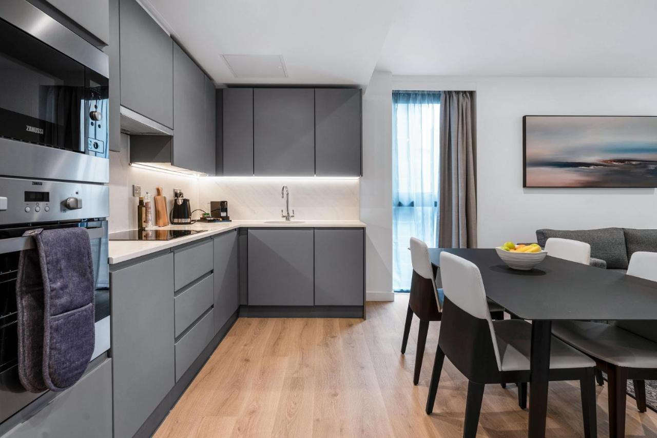 Citysuites 2 Aparthotel Σάλφορντ Εξωτερικό φωτογραφία The photo shows a modern kitchen and dining area. The kitchen features sleek gray cabinetry, a white countertop, and built-in appliances such as an oven. There is a sink and some kitchen utensils displayed. The adjacent dining area contains a black t
