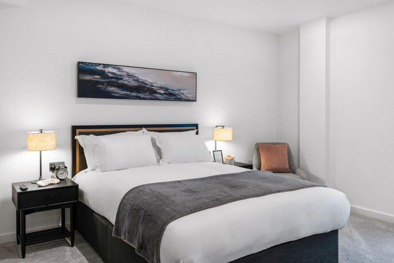 Citysuites 2 Aparthotel Μάντσεστερ Εξωτερικό φωτογραφία The photo shows a modern bedroom featuring a large bed with crisp white linens and a grey throw blanket. On either side of the bed, there are bedside tables with lamps. A piece of abstract art hangs above the headboard, and there is a chair in a corn