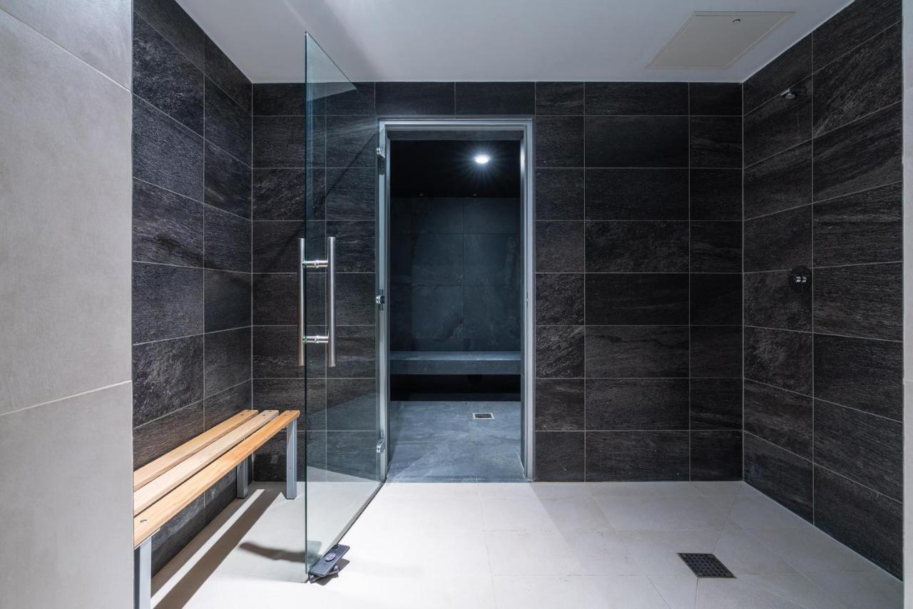 Citysuites 2 Aparthotel Μάντσεστερ Εξωτερικό φωτογραφία The photo shows a modern, minimalist shower area. The space is predominantly tiled with dark gray tiles on the walls, giving it a sleek and sophisticated look. There is a glass door that leads to another room, and a wooden bench is positioned against