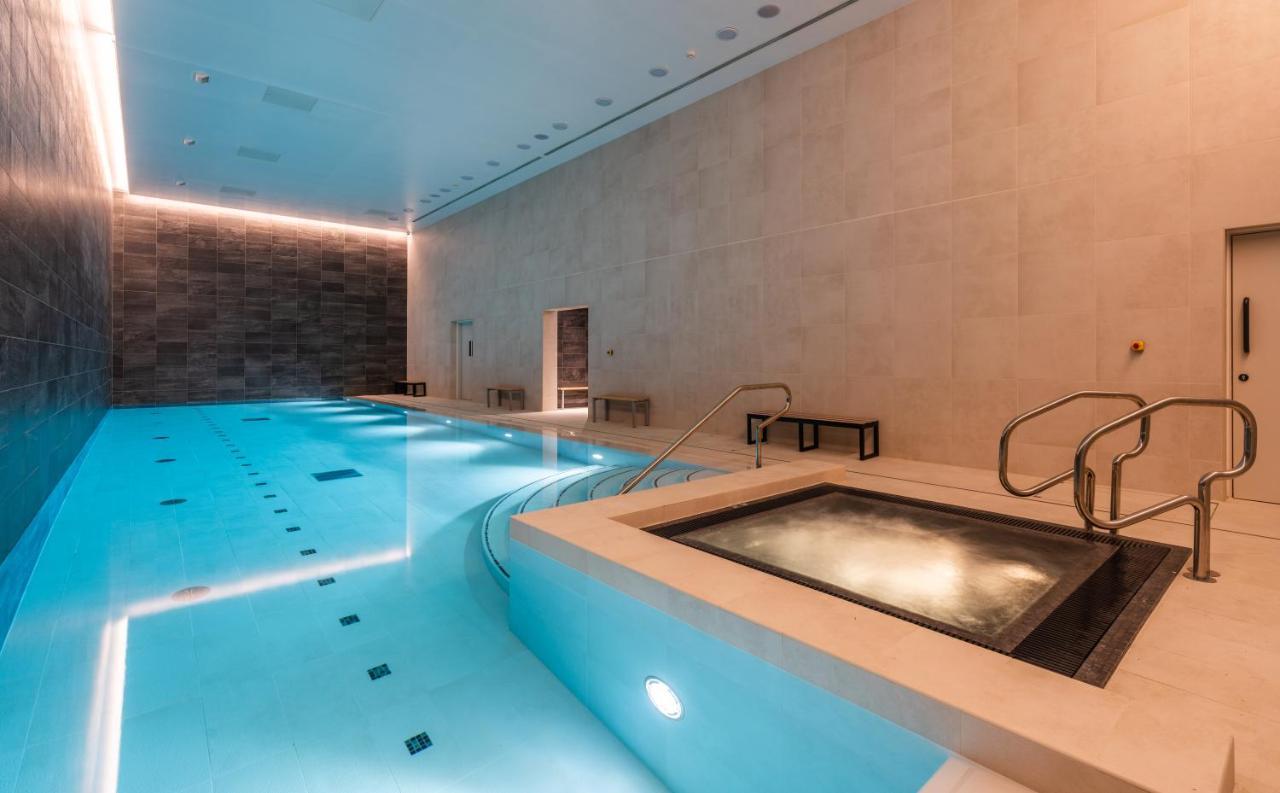 Citysuites 2 Aparthotel Μάντσεστερ Εξωτερικό φωτογραφία The photo shows an indoor swimming pool area. The pool is a bright blue color and features smooth, tiled surfaces. On one side, there is a hot tub area with steps leading into it. The walls are made of light-colored stone, and there is ambient lighti