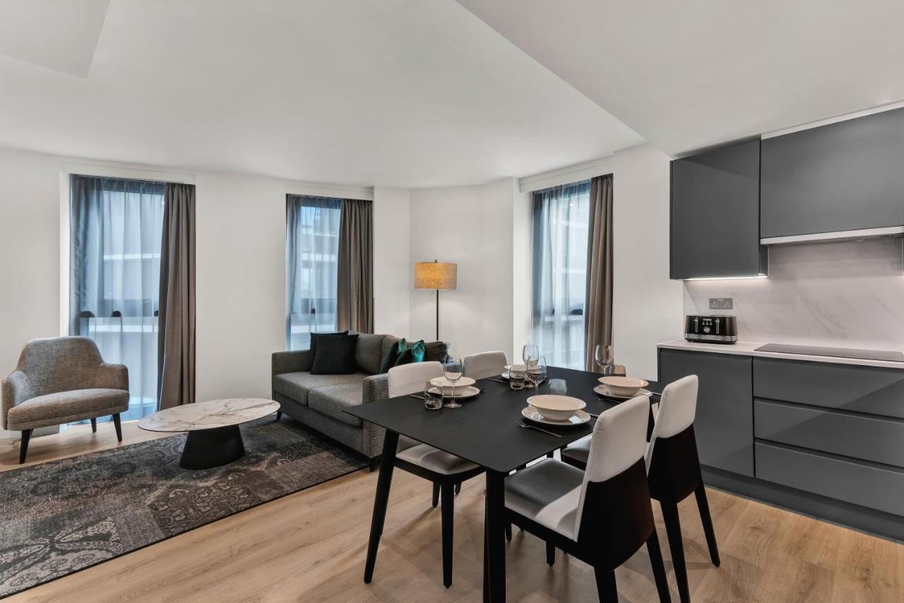 Citysuites 2 Aparthotel Μάντσεστερ Εξωτερικό φωτογραφία The photo shows a modern living space that includes a dining area and a seating area. There's a sleek black dining table set with plates and cutlery, accompanied by white and black chairs. To one side, a comfortable gray sofa faces the dining area, c