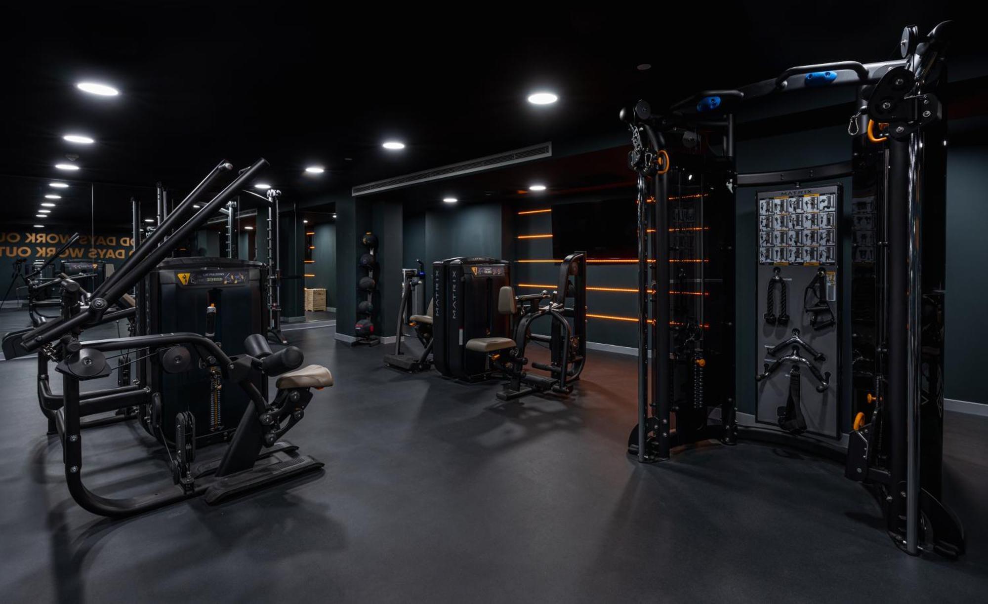 Citysuites 2 Aparthotel Μάντσεστερ Εξωτερικό φωτογραφία The photo shows a modern gym or fitness area with various workout machines. The space has a dark color scheme, featuring black walls and flooring. There are multiple pieces of exercise equipment, including stations for strength training, possibly tar