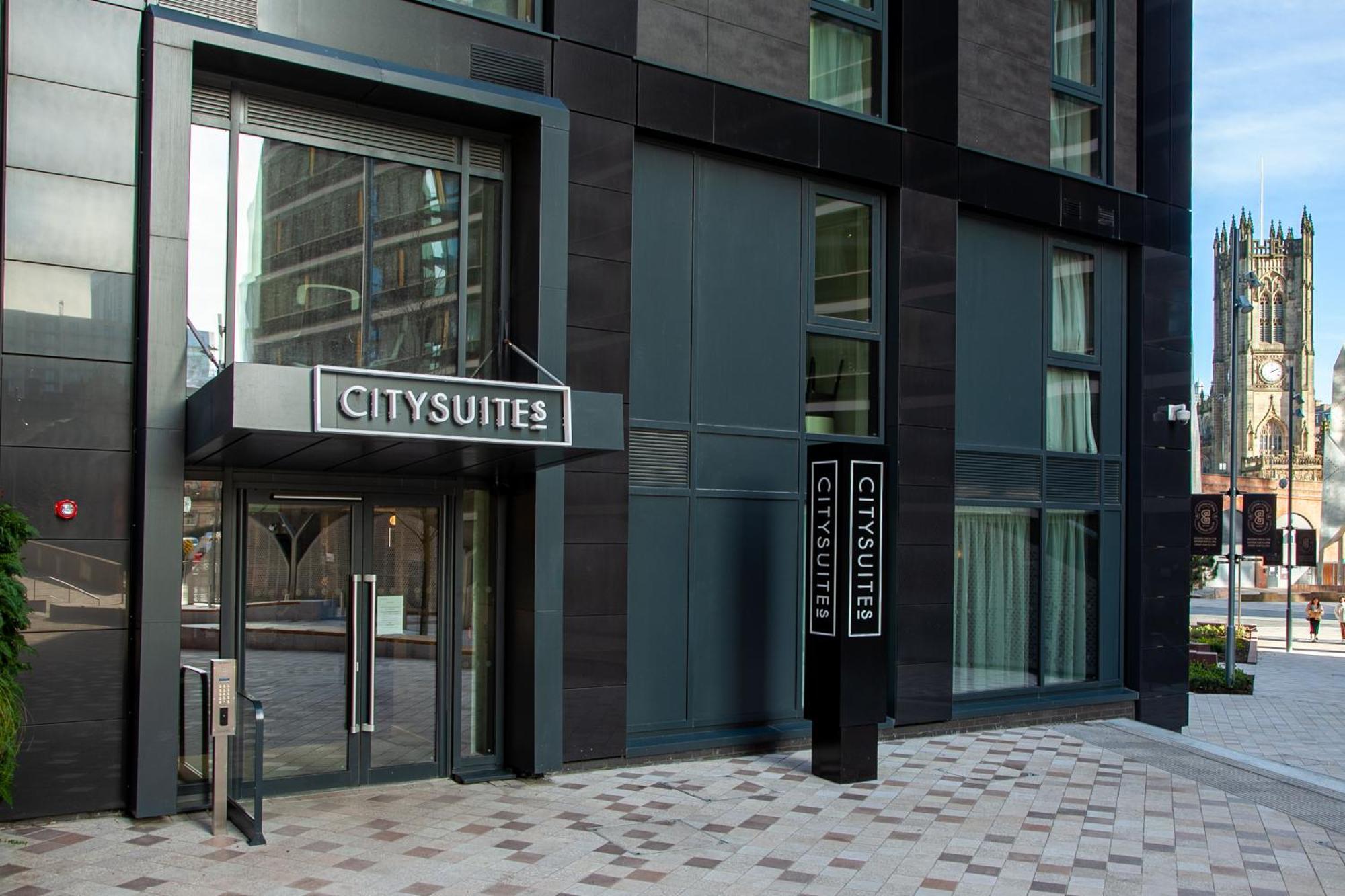 Citysuites 2 Aparthotel Μάντσεστερ Εξωτερικό φωτογραφία The photo shows the entrance to a modern building labeled "CITYSUITES." The entrance features large glass doors and a sleek, contemporary design. The building has dark exterior panels and large windows, suggesting a stylish and urban environment. The